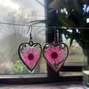May include: Heart-shaped earrings with bronze frames, each containing a pink flower preserved in clear resin. The earrings are suspended on a wire. The background is blurred, with a view of a window and foliage.