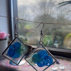 May include: Handmade earrings with blue pressed flowers set in clear resin within silver square frames. The earrings are suspended from silver-coloured fish hook ear wires, displaying a delicate floral design.
