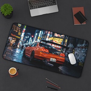 May include: A rectangular desk mat featuring an orange sports car driving down a neon-lit street in Japan. The mat includes a computer mouse, pencils, a coffee cup, and a small potted plant.