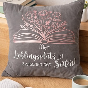 Könnte beinhalten: Graues Zierkissen mit einem rosa Blumendesign, das aus einem offenen Buch hervorgeht. Der Text "Mein Lieblingsplatz ist zwischen den Seiten!" ist in Weiß gedruckt. Ein Buch, eine Brille und eine Kaffeetasse sind ebenfalls zu sehen.