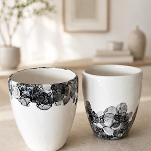 Könnte beinhalten: Zwei weiße Keramikbecher mit einem handgemalten schwarzen Tusche-Design. Der linke Becher zeigt das abstrakte, blumenartige schwarze Muster am Rand, während der rechte Becher dasselbe dunkle Tuschemuster im unteren Bereich aufweist.