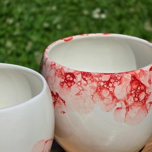 May include: Two white ceramic bowls with a rounded shape. The larger bowl features a vibrant red bubble pattern concentrated around the upper rim. The smaller bowl shows a faint red bubble design near the base. The text "tegla MERY" is visible.