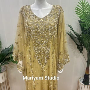 May include: A long, gold-colored dress with intricate beadwork and embellishments. The dress features a V-neckline and long, sheer sleeves. The text "Mariyam Studio" is visible on the dress. The dress is displayed on a mannequin.