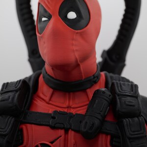 Peut inclure: Une figurine de Deadpool avec un masque rouge et noir aux yeux blancs, un plastron rouge orné d'un logo circulaire et un équipement tactique noir. Deux poignées d'épées noires dépassent de derrière les épaules du personnage.