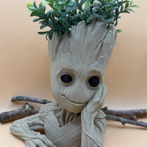 Peut inclure: Un pot de fleurs en forme de Groot de couleur beige avec des plantes artificielles vertes. Le pot a des yeux noirs et repose sa tête sur sa main. Deux petites branches sont derrière le pot.