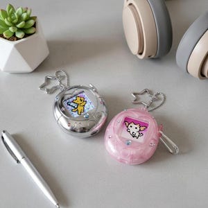May include: Two handheld digital pet devices, one silver and one pink, each with a pixelated character on the screen. The silver device has a star-shaped keychain. A succulent plant in a white geometric pot and a pen are also visible.