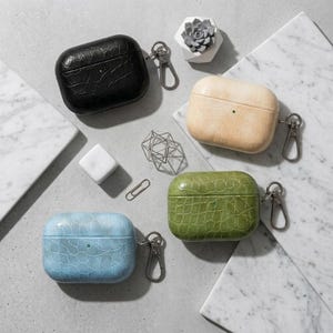May include: An assortment of four AirPod cases in black, beige, green, and blue, each with a crocodile skin texture. The cases have a silver carabiner clip attached. The items are arranged on a gray and white marble surface.