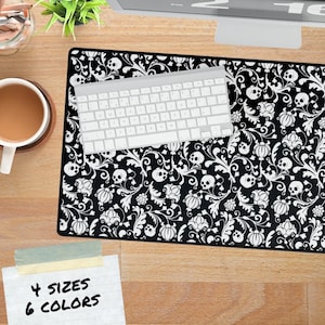May include: Black and white mouse pad with a repeating pattern of skulls and floral elements. A white keyboard rests on the pad. The image also shows a coffee cup, a plant, and a sticky note that says "4 sizes 6 colors."