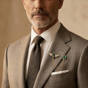 Peut inclure: Un costume gris avec une chemise blanche et une cravate marron. Une broche abeille dorée avec des accents verts et clairs est épinglée au revers, reliée par une chaîne à une broche carrée verte. Une pochette blanche est dans la poche poitrine.