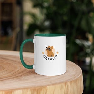 May include: White ceramic mug with a green handle and interior. The mug features a cartoon dog illustration with the text "LITTLE FECKER" in a playful font. The mug is sitting on a wooden surface.