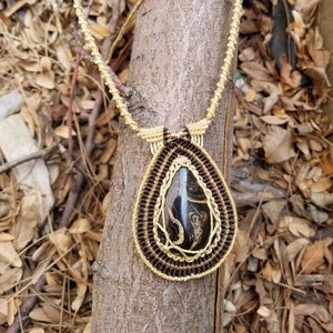 May include: A teardrop-shaped pendant necklace with a black and gold stone set in a macrame design. The necklace is made with brown and gold cord and is hanging from a tree branch.
