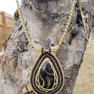 May include: A black and white marbled stone pendant hangs from a macrame necklace made with brown and beige cord. The necklace is draped over a tree trunk.