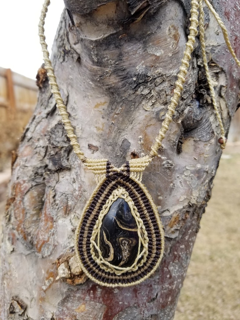 May include: A black and white marbled stone pendant hangs from a macrame necklace made with brown and beige cord. The necklace is draped over a tree trunk.