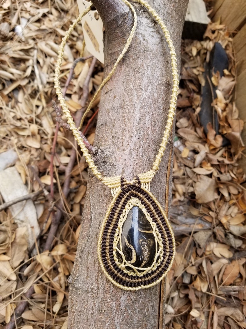 May include: A teardrop-shaped pendant necklace with a black and gold stone set in a macrame design. The necklace is made with brown and gold cord and is hanging from a tree branch.