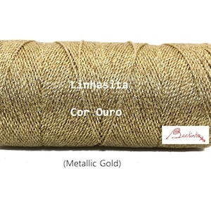 May include: A spool of metallic gold Linhasita cord. The cord is tightly wound and has a textured appearance. The text "Linhasita" and "Cor Ouro" are visible on the spool, along with the words "(Metallic Gold)". A logo is also present.