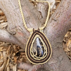 May include: A teardrop-shaped pendant necklace with a black and gold stone set in a macrame design. The macrame is made with brown and beige cord.