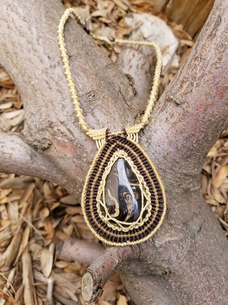 May include: A teardrop-shaped pendant necklace with a black and gold stone set in a macrame design. The macrame is made with brown and beige cord.