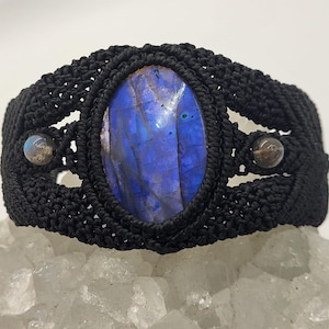 May include: A black macrame bracelet featuring an oval, iridescent blue labradorite stone. The bracelet has two smaller, round stones on either side of the central stone. The bracelet is displayed on a white, crystalline surface.