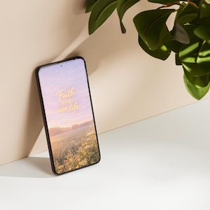 Puede incluir: Un smartphone con marco negro muestra un fondo de pantalla de paisaje con el texto "Faith brings new life" en dorado. Hojas verdes en la esquina superior derecha. El teléfono se apoya en una pared beige.