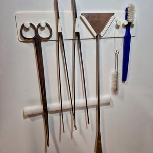 Puede incluir: Un conjunto de herramientas para acuarios, que incluye tijeras, pinzas, un raspador y un cepillo, montadas en un soporte blanco. Las herramientas son de metal y plástico. El cepillo tiene plástico azul y cerdas blancas.