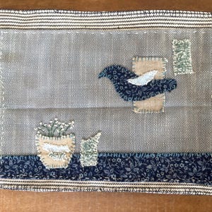 Op de afbeelding: Een rechthoekige stoffen applicatie met een grijze achtergrond, een donkerblauwe vogel met patroon, een beige pot met witte bloemen en kleine groene lapjes. Het stuk is omlijst door een gestreepte rand en een donkerblauwe strook bloemenstof.