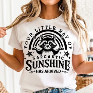 K&ouml;nnte beinhalten: Wei&szlig;es T-Shirt mit einer schwarz-wei&szlig;en Waschb&auml;r-Grafik und dem Text "YOUR LITTLE RAY OF SARCASTIC SUNSHINE HAS ARRIVED". Das Design beinhaltet einen Regenbogen, Sterne und dekorative Pfeile.