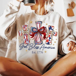 Può includere: Una felpa a maniche lunghe color crema con una grafica di tre croci decorate con motivi e fiocchi rossi, bianchi e blu. Il testo recita God Bless America ed Est. 1776, circondato da piccole stelle dorate sulla parte anteriore del capo.