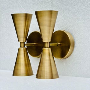 Op de afbeelding: Twee messing wandkandelaars. Elke wandlamp heeft een conische kap boven een kleinere, omgekeerde conische basis, verbonden door een slanke staaf. De wandkandelaars zijn gemonteerd op ronde achterplaten. De afwerking heeft een warme, antieke uitstraling.