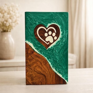 Könnte beinhalten: Rechteckige Urne für Haustiere aus Holz mit herzförmigem Ausschnitt und Pfotenabdruck. Die Urne hat eine teal-farbene Harzoberseite und einen Holzsockel. Das Design umfasst einen weißen Akzent entlang der Holzmaserung.