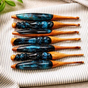 Peut inclure: Six crochets en bois sont présentés. Les manches combinent bois et résine, la résine présentant un motif tourbillonnant bleu et noir. Les crochets sont disposés sur un tissu rayé, soulignant leur savoir-faire et leur design.