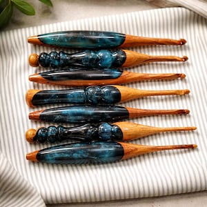 May include: Six crochet hooks with wooden handles and dark blue and black resin accents. The hooks are displayed on a striped fabric. The design highlights the contrast between the wood and resin, showcasing the tools.