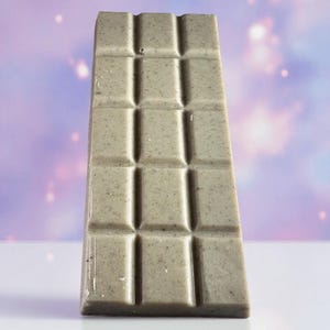 May include: A rectangular bar of light grey, speckled solid material divided into twelve equal segments by a grid of indented lines. The surface has a smooth, matte texture with small dark flecks distributed throughout the entire structure.