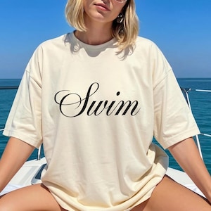 May include: Cream-colored t-shirt with the word "Swim" in elegant black script. The shirt is a relaxed fit, perfect for a day at the beach or pool. The background shows a clear blue sky and water.