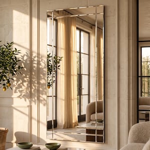 May include: A tall, rectangular wall mirror featuring a segmented frame with thin, metallic silver-toned dividers. The mirror reflects a bright interior space with sheer curtains, a glass door, and a portion of a beige upholstered chair.