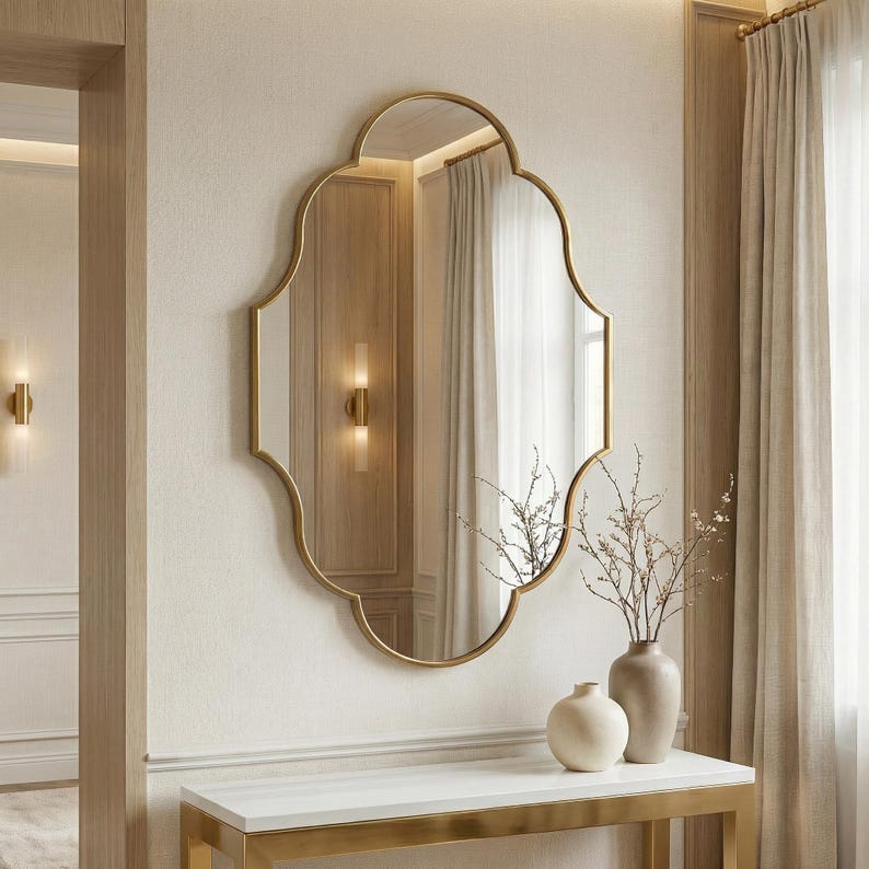 May include: A gold-framed mirror with an ornate, scalloped shape hangs on a cream-colored wall. The mirror reflects a room with a window and curtains. A white console table with gold trim is below the mirror, with vases.