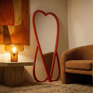 May include: A modern interior scene featuring a heart-shaped red mirror, a brown armchair, and a table with a mushroom-shaped lamp. The lamp is amber-colored and sits on a square, light-colored stone table. An abstract painting hangs on the wall.