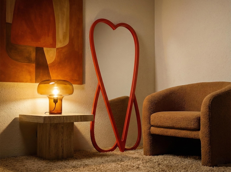 May include: A modern interior scene featuring a heart-shaped red mirror, a brown armchair, and a table with a mushroom-shaped lamp. The lamp is amber-colored and sits on a square, light-colored stone table. An abstract painting hangs on the wall.