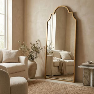 Puede incluir: Un espejo alto con marco dorado y parte superior arqueada en una habitaci&oacute;n de tonos neutros. El espejo refleja un sof&aacute;, cojines y una alfombra. El marco tiene una forma decorativa. La habitaci&oacute;n tiene un esquema de color beige.