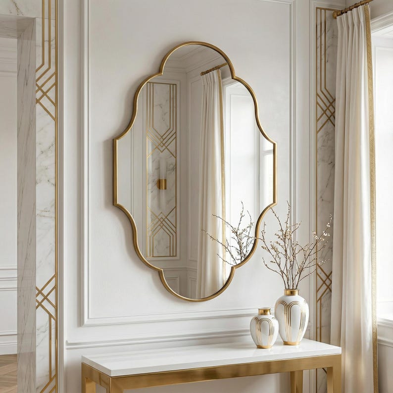 May include: A gold-framed wall mirror with an ornate, scalloped shape hangs above a white console table. The mirror reflects a room with white walls, gold accents, and a window with white curtains. Two decorative vases sit on the table.