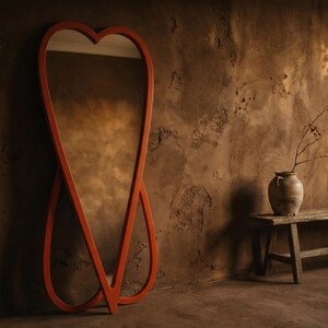 May include: A large, orange, heart-shaped full-length mirror leans against a textured brown wall. The mirror's frame has a unique, intertwined design. A rustic bench and vase with branches are in the background, creating a warm, inviting atmosphere.
