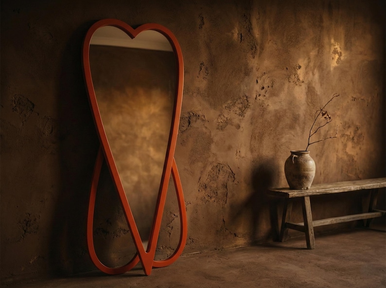 May include: A large, orange, heart-shaped full-length mirror leans against a textured brown wall. The mirror's frame has a unique, intertwined design. A rustic bench and vase with branches are in the background, creating a warm, inviting atmosphere.