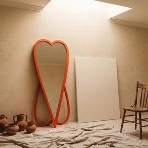 May include: An orange heart-shaped mirror leans against a beige wall, next to a wooden chair and a blank white canvas. Several terracotta pots and a draped cloth are on the floor. The room is lit by natural light.