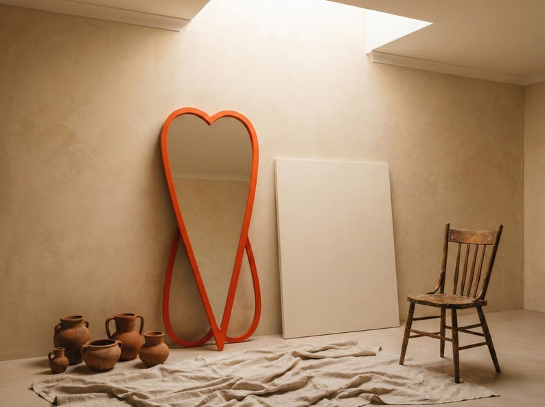 May include: An orange heart-shaped mirror leans against a beige wall, next to a wooden chair and a blank white canvas. Several terracotta pots and a draped cloth are on the floor. The room is lit by natural light.