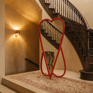 May include: A red heart-shaped full-length mirror stands on a patterned rug in a hallway. The mirror's frame is a bold red, and the design is a continuous loop. A wooden staircase and a small table are visible in the background.