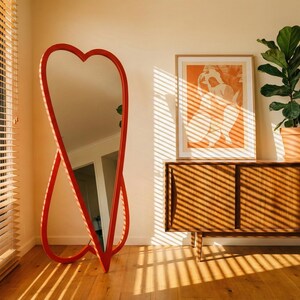 May include: A red heart-shaped floor mirror stands in a room with a wooden cabinet, a framed print, and a potted plant. The mirror's frame is a vibrant red, and the room is bathed in sunlight from a window with blinds.