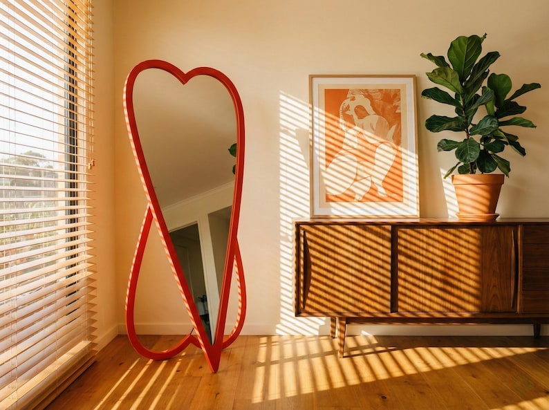May include: A red heart-shaped floor mirror stands in a room with a wooden cabinet, a framed print, and a potted plant. The mirror's frame is a vibrant red, and the room is bathed in sunlight from a window with blinds.