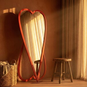 May include: A large, red heart-shaped mirror stands against a warm-toned wall. A woven basket and a wooden stool are nearby. The mirror reflects a window with sheer curtains, creating a soft, inviting atmosphere. The room is bathed in warm sunlight.