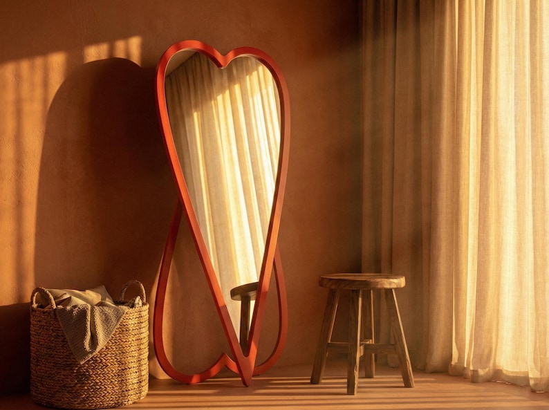 May include: A large, red heart-shaped mirror stands against a warm-toned wall. A woven basket and a wooden stool are nearby. The mirror reflects a window with sheer curtains, creating a soft, inviting atmosphere. The room is bathed in warm sunlight.