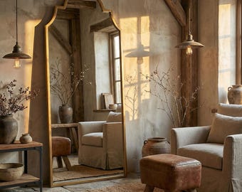 May include: A tall, gold-framed mirror with an arched top stands in a rustic room. The mirror reflects the room's interior, including two beige armchairs, a leather ottoman, and decorative vases. The room has a warm, inviting atmosphere.