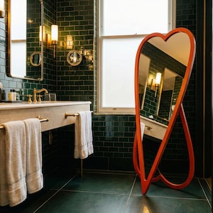May include: A bathroom with dark green tile walls and a matching floor. A large, orange, heart-shaped mirror stands against the wall. A white countertop with gold fixtures and a window are also visible.